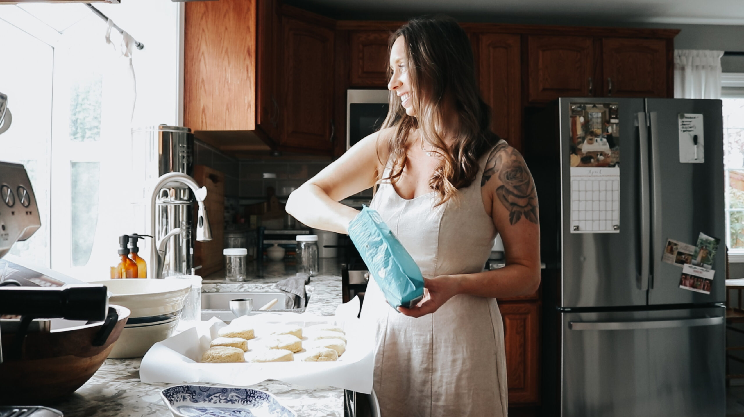 Christian homemaker stands at her kitchen counter in front of a window with her hand in a bag of flour about to sprinkle it on uncooked biscuits
